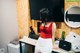 A woman in a red top and white skirt standing in front of a desk.